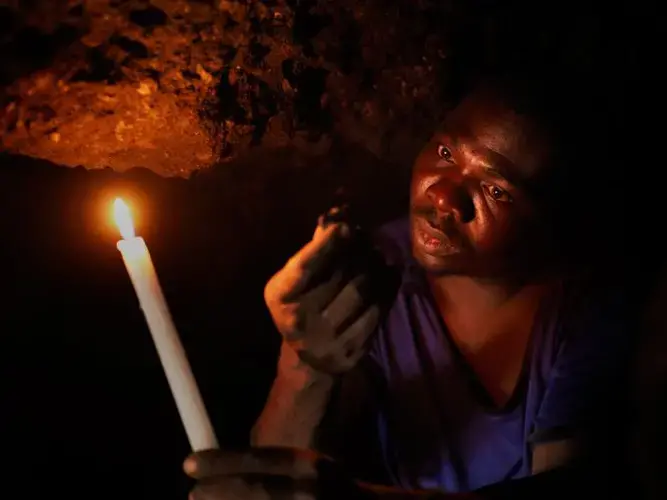 Deep inside a horizontal shaft near the top of Black Mountain, a miner uses a candle to illuminate a small piece of lead ore. Miners look for brown slag, which contains the highest concentrations of lead. Tunneling through the soft tailings is dangerous. No support are used to shore the tunnel roof and walls and collapses sometimes occur. Over the years, many miners have lost their lives working on the mountain. Image by Larry C. Price. Zambia, 2017.