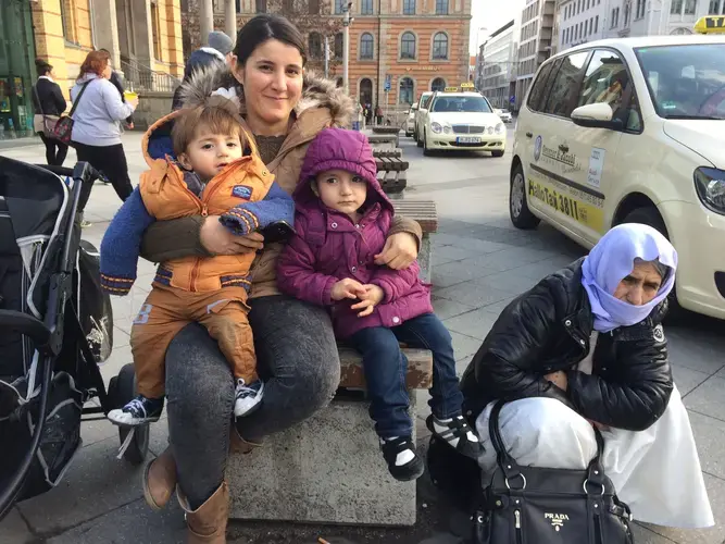 Amsha Ali Aylas, accompanied by her mother-in-law Khunaf, holds her two children outside of a train station in Germany. Image by Emily Feldman. Germany, 2016.