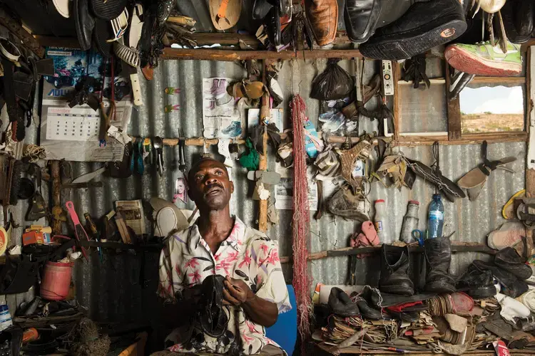 Eddy Bien Aime fixes a pair of sandals at his business in the neighborhood known as Canaan 2. Most of his business involves selling footwear he has bought secondhandand refashioned. He moved to Canaan just days after the earthquake and claimed a large parcel of land, some of which he has since given away. Image by Allison Shelley. Haiti, 2017.<br />
