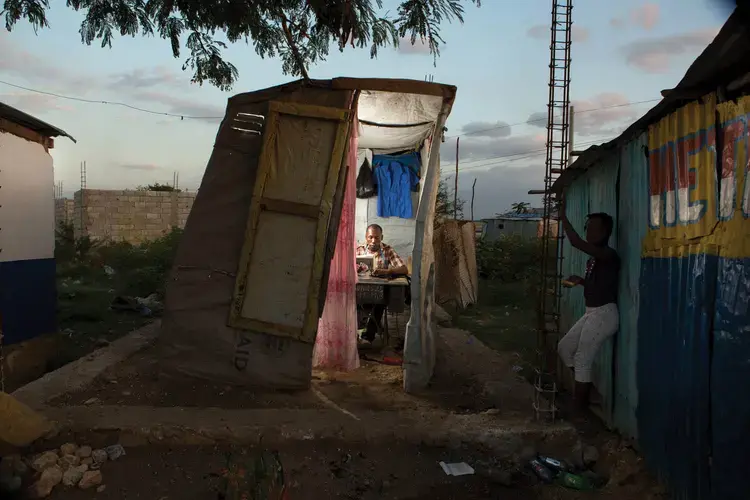 Reginald Adrasse repairs a uniform in his small self-built tailor shop at the entrance to Canaan 2. Image by Allison Shelley. Haiti, 2017.