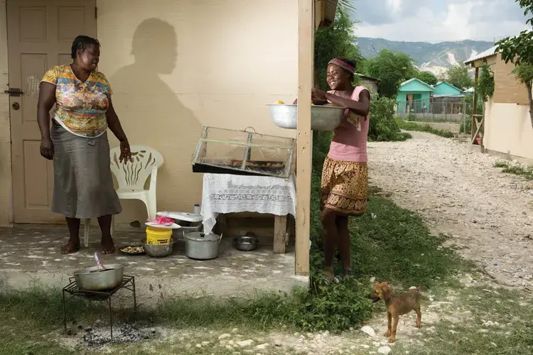 Pascal Amelina (left) and her neighbor sell fritay—fried snacks—in the Corail section of Canaan. Amelina has a handful of small businesses that sell wine, embroidered clothing, sweets, and other items. Image by Allison Shelley. Haiti, 2017.<br />
