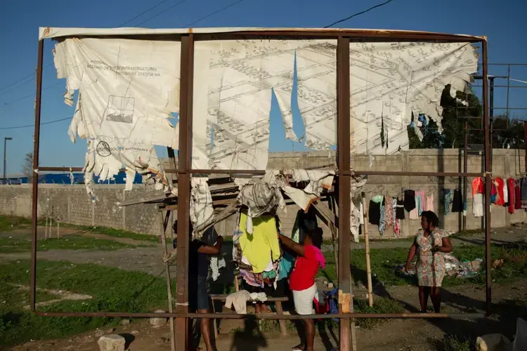 Women sell secondhand clothing beneath the tatters of a sign that once promoted the Caracol Industrial Park public funding agreement along with a rendering of its grounds, at the main entrance to the park, in Caracol, Haiti, December 9, 2019. Image by Allison Shelley. Haiti, 2019.