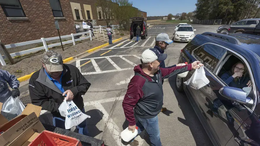 Future Farmers of America Alumni including Dennis Roehl, left, Adam Luchterhand and Tom Odeen hand out free food on May 7, 2020 outside the public schools in Loyal, Wis. Each family who pulled up received a gallon of milk, cheese, butter and beef sticks. At the same time the school was distributing their daily free lunches to anyone under the age of 18. Image by Mark Hoffman / Milwaukee Journal Sentinel. United States, 2020.