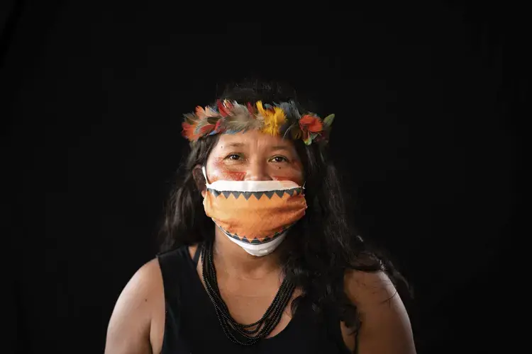 Terezinha de Souza, 45, of the Sateré Mawé indigenous ethnic group, poses for a portrait wearing the traditional dress of her tribe and a face mask amid the spread of the new coronavirus in the Gaviao community near Manaus, Brazil, Friday, May 29, 2020. At the end of May, de Souza fell ill with fever, body aches, and a dry cough, and when her health deteriorated she crossed the river to the indigenous wing of the Manaus hospital, inaugurated just a few days earlier, where she has first turned away but later admitted. In de Souza's opinion, hospitalizations in her community could have been avoided had a nurse or doctor visited them at earlier stages of the pandemic. Image by Felipe Dana / AP Photo. Brazil, 2020.