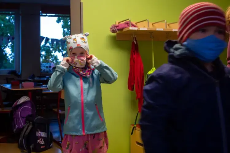 A young student dons a mask after putting on rain gear to go outside for recess at a primary school in Weimar, Germany. Students are only required to wear their masks when walking through hallways, though tighter measures could be put in place if coronavirus cases in Weimar increase. Image by Ryan Delaney/St. Louis Public Radio. Germany, 2020.