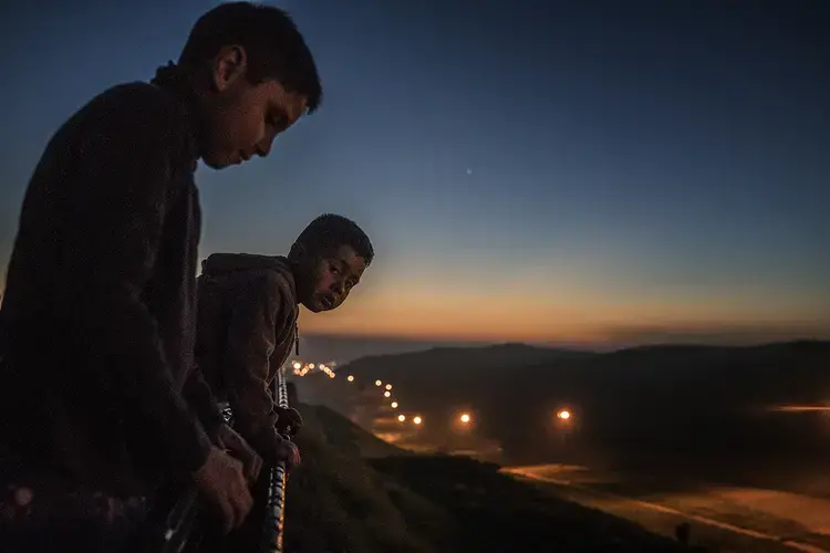 Young boys from the Arias family peer over the first, older U.S.-built border wall at dusk into the flood-lit no-man's land created between the two walls/fences where U.S. Border Patrol is present 24 hours a day, seven days a week. Image by James Whitlow Delano. Mexico, 2017.<br />