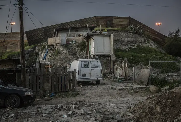 Flood lights from the U.S. side of the border begin to bear down on a house in Otay Canyon (Cañon de Otay) that rests up against the border wall, as night falls on the U.S./Mexico Border. Image by James Whitlow Delano. Mexico, 2017.