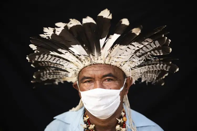 Indigenous leader Pedro dos Santos, of the Mura indigenous ethnic group, poses for a portrait wearing the traditional dress of his tribe and a face mask amid the spread of the new coronavirus in the Park of Indigenous Nations community in Manaus, Brazil, Thursday, May 28, 2020. In April, dos Santos, his wife, children, and grandchildren all fell ill with COVID-19 symptoms. After days of fever, the 70-year-old chief sought treatment in a public clinic, but the nurse said he’d be better off staying at home, so he returned without ever being tested for the new coronavirus. Some of his family members are still sick and recovering at home. Image by Felipe Dana / AP Photo. Brazil, 2020.