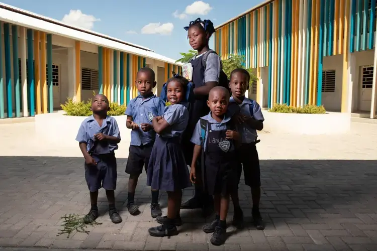 Fifth grade student Kerlande Vezilord, 16, poses for a photo with kindergarteners at Ecole Fondamentale Complete Breda de Canaan, greater Canaan's very first public school. Like every student, Vezilord scored at the top of a competitive entrance exam in order to enroll. She wakes up at 4 a.m. to cook for her family and leaves home at 6 a.m. every weekday for her hour-long walk to class. 'Education is important because one day it will allow me to speak your language with you,' says Vezilord, whose dream is to become a nurse in either the U.S. or Cuba. Image by Allison Shelley. Haiti, 2019.