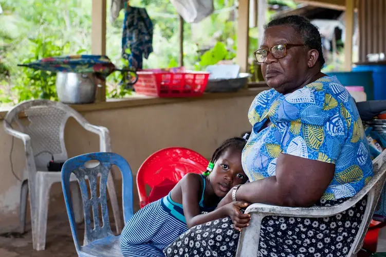 Mercy Prika, 5, lays her head in the lap of her grandmother, village Captain Wilma Prika, as she sits at her house in Adjuma Kondre, Suriname. “They made a lot of money from this mountain, and they didn’t do anything for the village,” said Captain Prika of Alcoa subsidiary Suralco's impact on her village after years of bauxite mining in the hills surrounding it. “I am actually ashamed that this is my village.” Image by Stephanie Strasburg. Suriname, 2017.