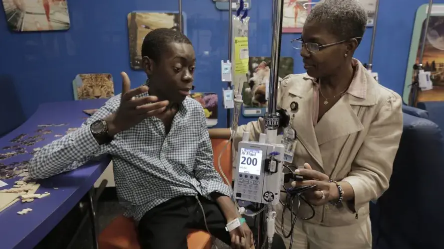Djonsly Alcin (left), 14, talks to Baptist Health nurse Martha Pearson (right) during chemotherapy. Djonsly, who is from Haiti, suffers from optic pathway glioma, a slow-growing brain tumor. His advanced cancer has also spread down his spine. Image by José A. Iglesias. United States, 2018.