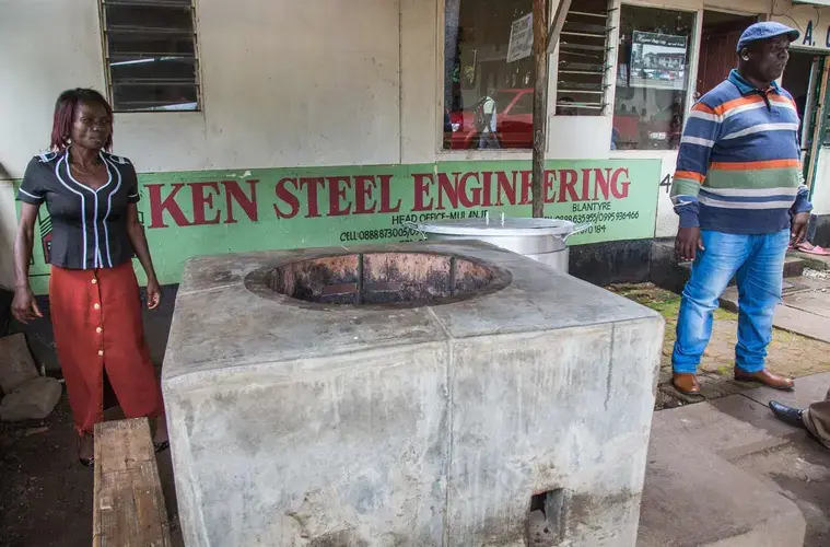 Ken Chilewe (right) is the most successful man in Mulanje – his business, Ken Steel Engineering, makes millions of dollars selling cookstoves. In front of his shop he built a rocket stove intended for serious cooks: the stove can hold a large pot for water; enough for the whole family. Image by Nathalie Bertrams. Malawi, 2017.