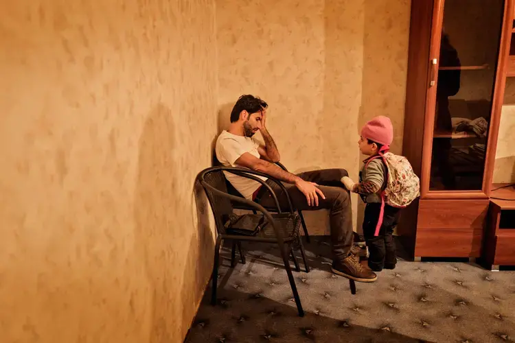 Syrian refugee Muhanned Abzali sits, exhausted, as his son Wael, stands beside him moments after arriving for the first time into the family's new apartment after a long, grueling day of travel and upheaval once again from Athens to their new home in Polva, Estonia. Image by Lynsey Addario. Estonia, 2017.