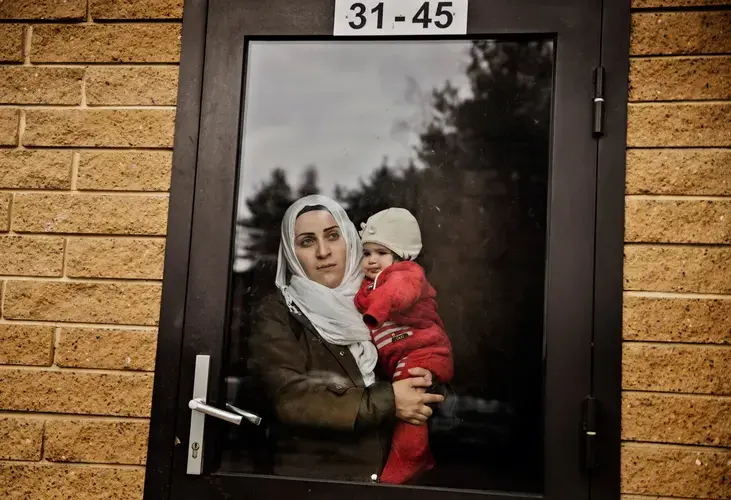 Syrian refugee Taima Abzali stares out the window of her apartment building front door as she holds baby Heln the morning after a long, grueling day of travel and upheaval once again from Athens to their new home in Polva, Estonia. Image by Lynsey Addario. Estonia, 2017.