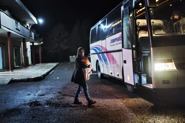 Syrian refugee Taimaa Abazli, 24, carries baby Heln, 4 months old, to a bus sponsored by UNHCR which will take refugee families to Athens for asylum appointments in Athens. Image by Lynsey Addario. Greece, 2017.