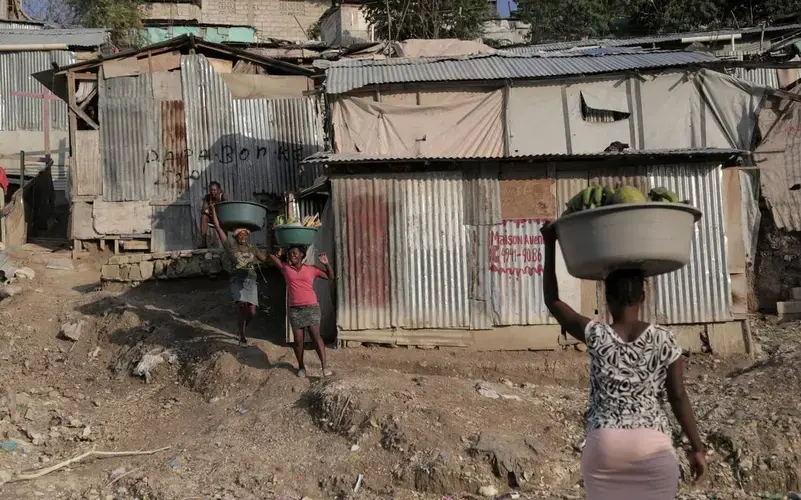 Women carrying produce and other goods walk by some of the tin shacks that make up the Teren Toto camp in Haiti’s capital. Image by Jose A. Iglesias. Haiti, 2019.