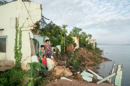 Ha Thi Be and her two grandsons stand in the ruins of her ancestral home along the Tien River in Dong Thap Province. The government warned her to relocate inland to live with her son in January 2017, two months before the house eventually collapsed. 'But everything I own was in this house, and now everything is gone,' she said. Image by Sim Chi Yin. Vietnam, 2017.