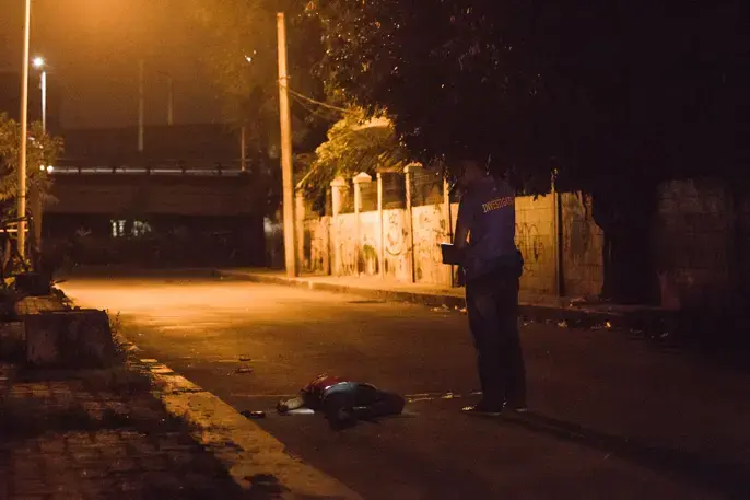 An investigator inspects the body of alias “John Rex,” who was on the village’s drug watch list. Police said he fell from a motorcycle that was driven by an unidentified companion while “John Rex” was shooting at the police. In what police said was an act of self-defense, they fired back at him. Image by Pat Nabong. Philippines, 2017.