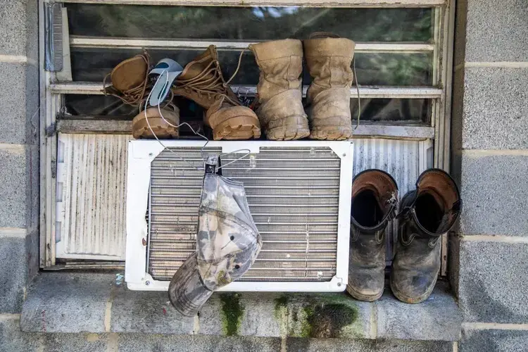 Boots and a hat dry on a window air conditioning unit at a Johnston County farmworker camp Thursday August 27, 2020. Air conditioning is considered a luxury by many farmworkers in North Carolina farmworker camps. Image by Travis Long. United States, 2020.