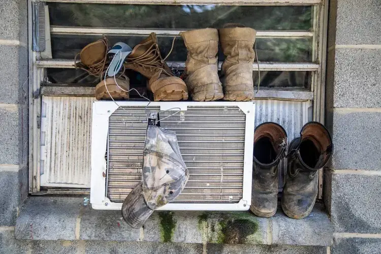 Boots and a hat dry on a window air conditioning unit at a Johnston County farmworker camp Thursday August 27, 2020. Air conditioning is considered a luxury by many farmworkers in North Carolina farmworker camps. Image by Travis Long. United States, 2020.