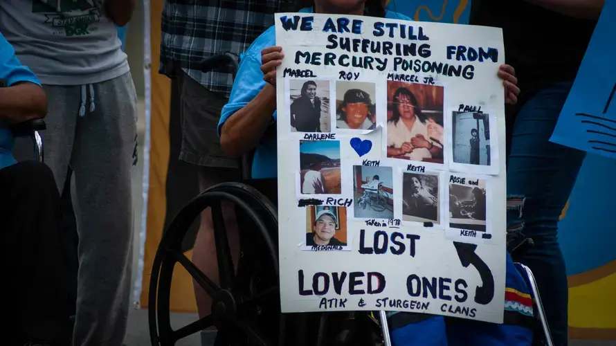 A woman sits in a wheelchair and holds a sign which reads, 'We are still suffering from mercury poisoning.' It also features photos of family members who have passed away.