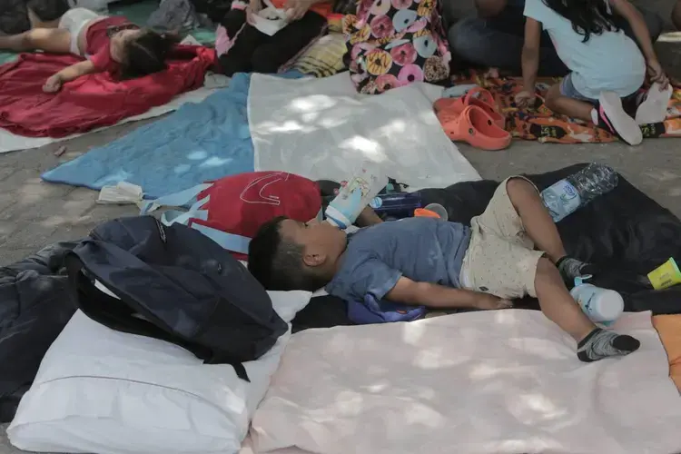 A young Peruvian boy drinks milk in the shade in a camp in a small park by the entrance to Gateway International Bridge in Matamoros, Mexico. Gateway International Bridge is one of three international bridges that cross the U.S.-Mexico border between the cities of Brownsville, Texas, and Matamoros, Tamaulipas. Image by Jose A. Iglesias. Mexico, 2019.