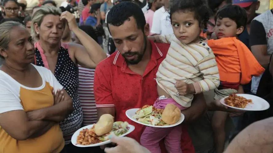 A migrant father and his daughter receive food at a migrant camp on the Matamoros side of the Gateway International Bridge that connects Matamoros, Mexico, to Brownsville, Texas. Church and volunteer organizations in Brownsville provide water and food for the migrants on the Mexican side of the border as they wait for their asylum requests to be processed. Image by Jose A. Iglesias. Mexico, 2019.