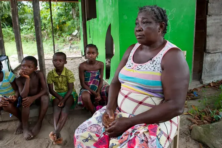 Emelien Adjako sits for an interview in her village of Kajapaati along the Suriname River. Adjako was one of those from over 40 Saamaka villages displaced by the flood waters caused by the formation of a reservoir with the construction of the Afobaka Dam. Built by Suralco to power a now defunct aluminum smelter, the dam now provides about half of Surinam's electricity, none of which has reached Adjako's replacement home. Image by Stephanie Strasburg/Pittsburgh Post-Gazette. Suriname, 2017.