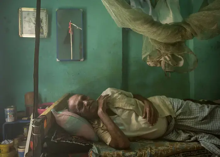 75-year-old Charira Sahni suffers from a chronic but undiagnosed lung condition. He wheezes and coughs relentlessly and spends much of his time in a bed tucked away in a tiny room off the dirt patio where his wife cooks. Image by Larry C. Price. India, 2018.