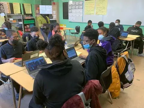 Black students, ages 12-13, in dark blue uniforms and wearing masks turn towards each other to have a discussion