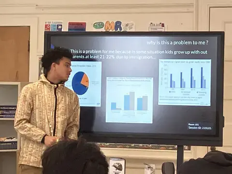 A teen stands in front of a screen and presents his presentation on immigration by displaying a slide with several graphs