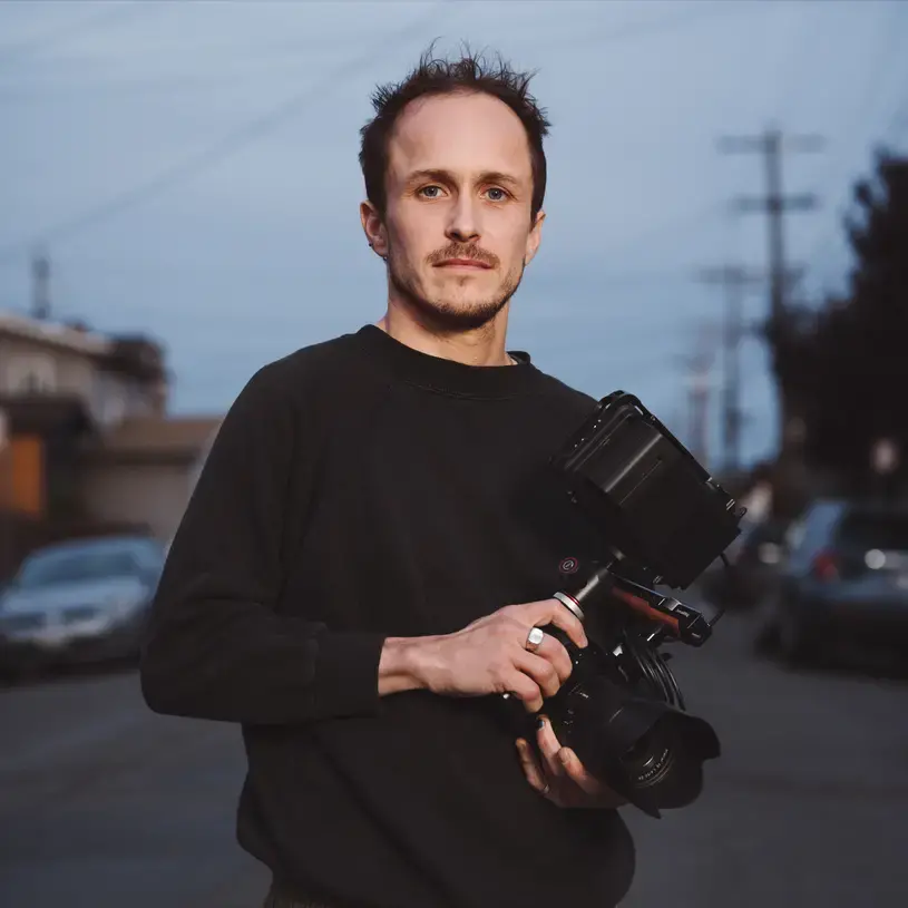 Brandon Tauszik Headshot of Brandon Tauszik. He wears a black long-sleeved crewneck sweatshirt and holds a video camera.