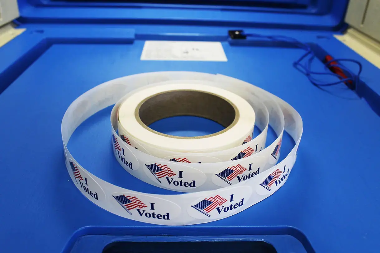 Red white and blue "I voted" stickers with American flag are on a spool of stickers placed on a voting booth platform. Image by Barbara Kalbfleisch. United States, undated. Red white and blue "I voted" stickers with American flag are on a spool of stickers placed on a voting booth platform. Image by Barbara Kalbfleisch. United States, undated.