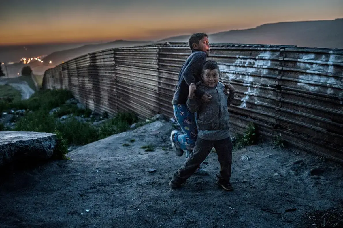 Two boys from the Arias family roughhouse in the dust beside their home abutting the original U.S.-built border wall in Colonia Libertad, Tijuana, Mexico. Behind them, a string of lights set up by U.S. Border Patrol stretches to the sea. The lights were set up by U.S. Border Patrol to illuminate the wall to make it more difficult migrants to cross at night. Image by James Whitlow Delano. Mexico, 2017. Two boys from the Arias family roughhouse in the dust beside their home abutting the original U.S.-built border wall in Colonia Libertad, Tijuana, Mexico. Behind them, a string of lights set up by U.S. Border Patrol stretches to the sea. The lights were set up by U.S. Border Patrol to illuminate the wall to make it more difficult migrants to cross at night. Image by James Whitlow Delano. Mexico, 2017.