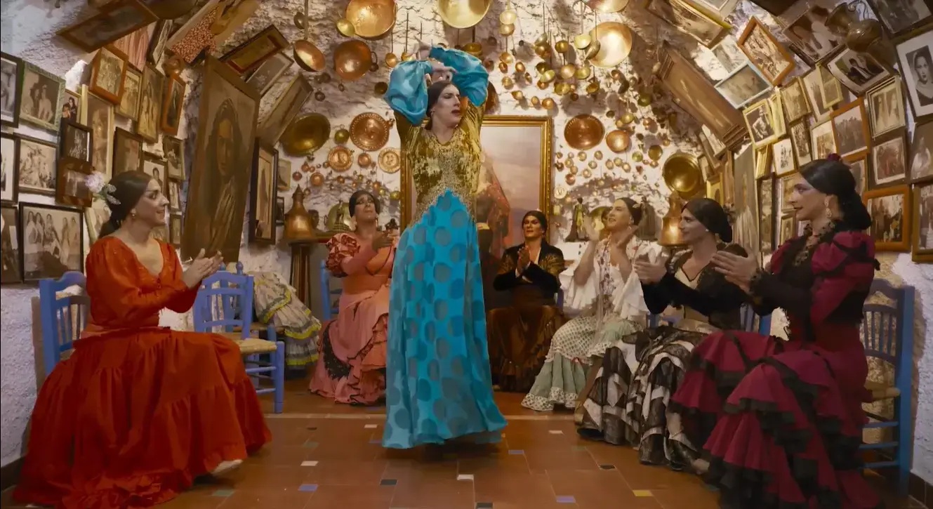 A man in drag wearing a blue and gold dress dances in the middle of a room. Sitting around the perimeter of the room and clapping along are five other men in drag. The walls are covered in gold decorations and framed pictures.