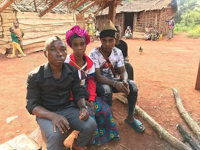 Three Baka individuals sit on a bench outside a wood and clay building