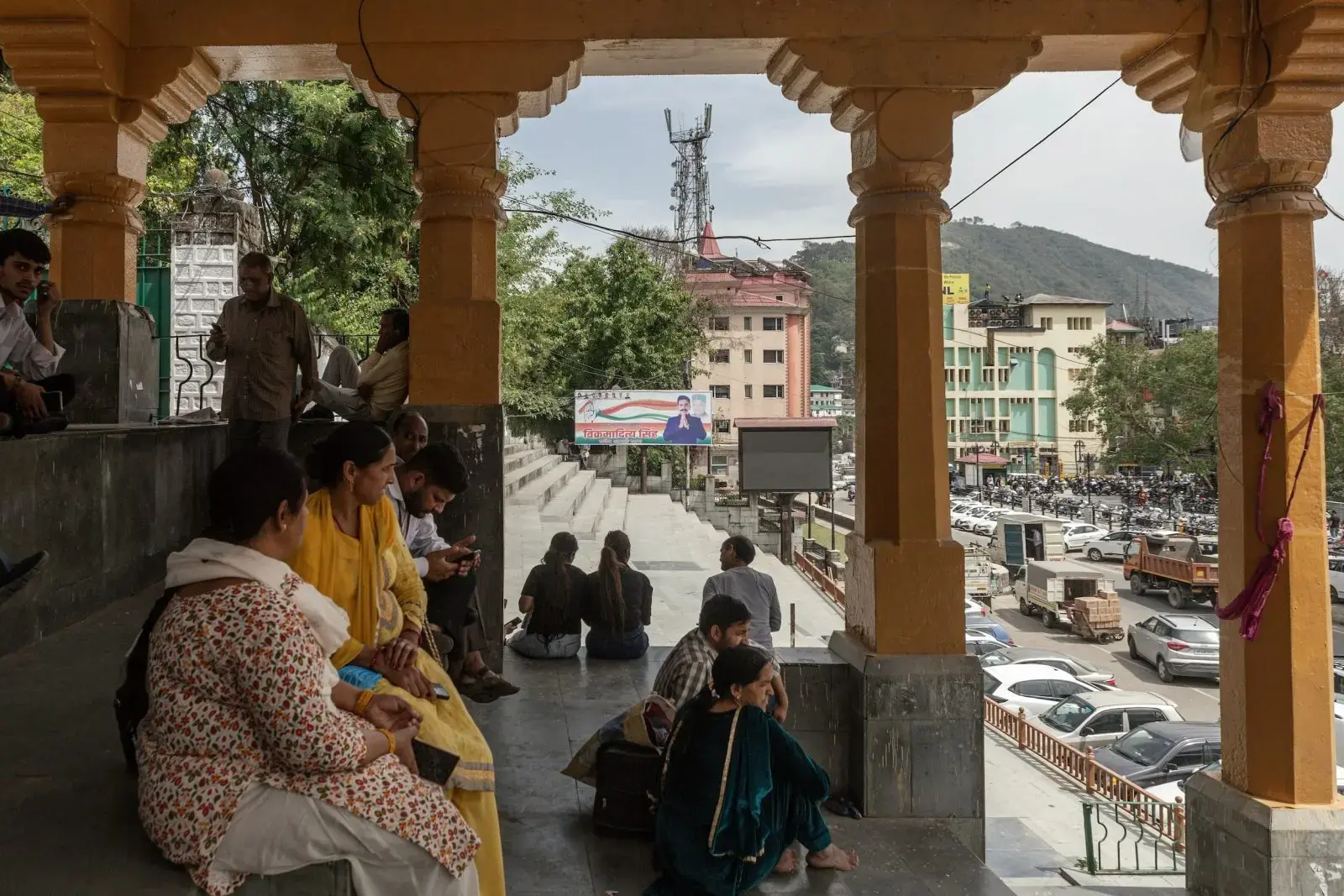 People sit on shaded stone steps lined by adorned columns, a cityscape in the background.