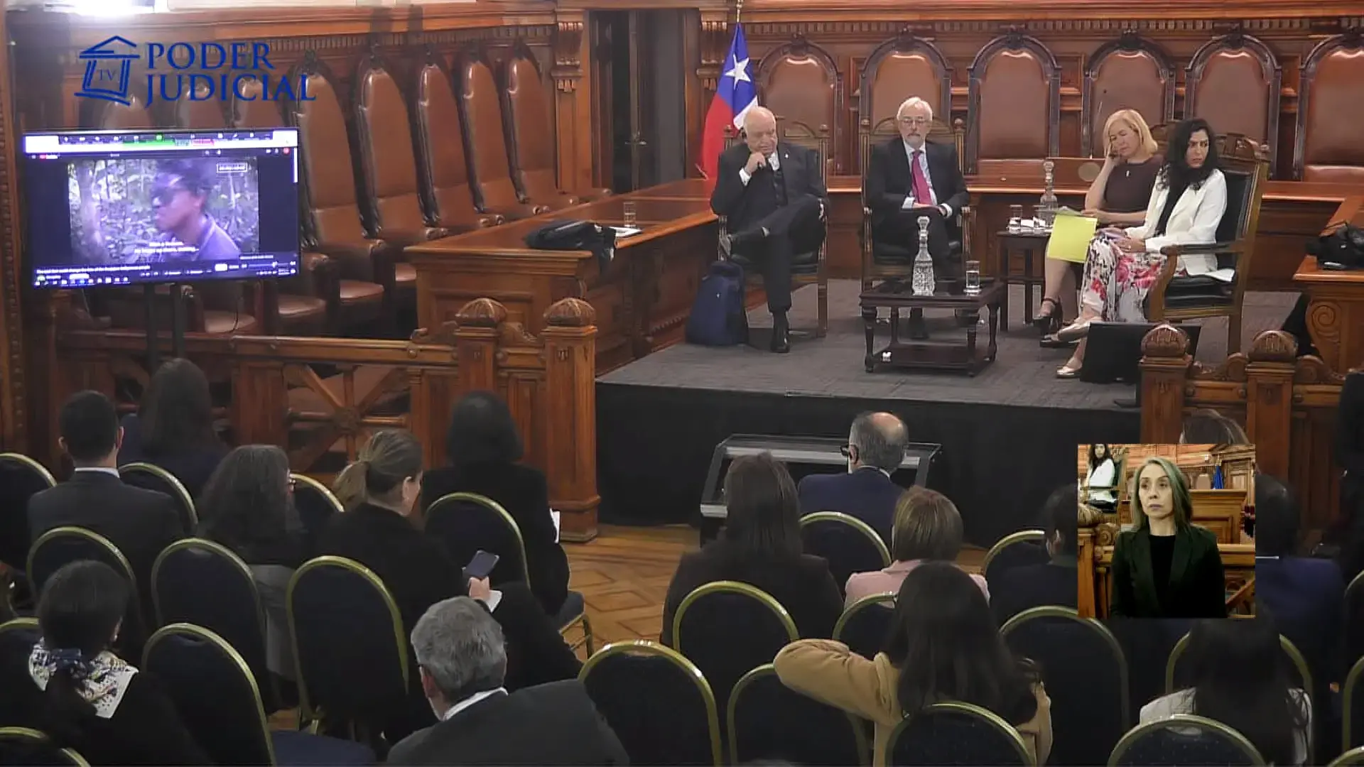 Four panelists sit on a stage wedged between rows of ornate seats lining the walls of a wooden-paneled room. They have an audience sitting in front of them and a television showing a video.