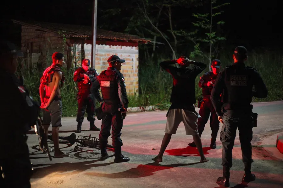 At night, lit by red lights, two men are detained on the roadside as several policemen stand by.