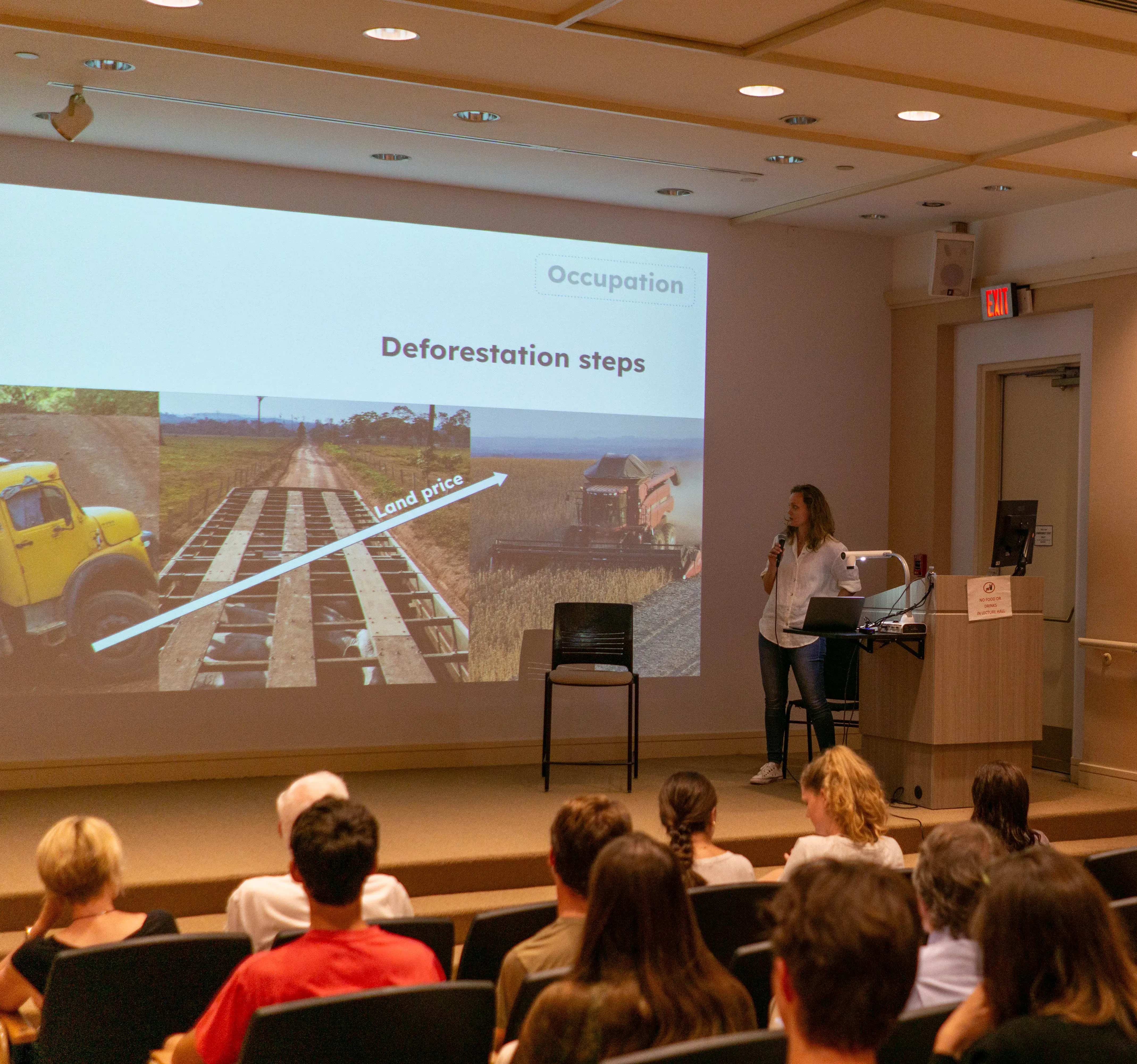 Fernanda stands in a lecture hall holding a microphone as she looks towards a projection of a presentation titled 'deforestation steps.' the pictures show a truck carrying lumber, a dirt road, and a soy combine, as a diagonal line goes up and right titled 'land price' indicating the value of the deforested land is increasing.