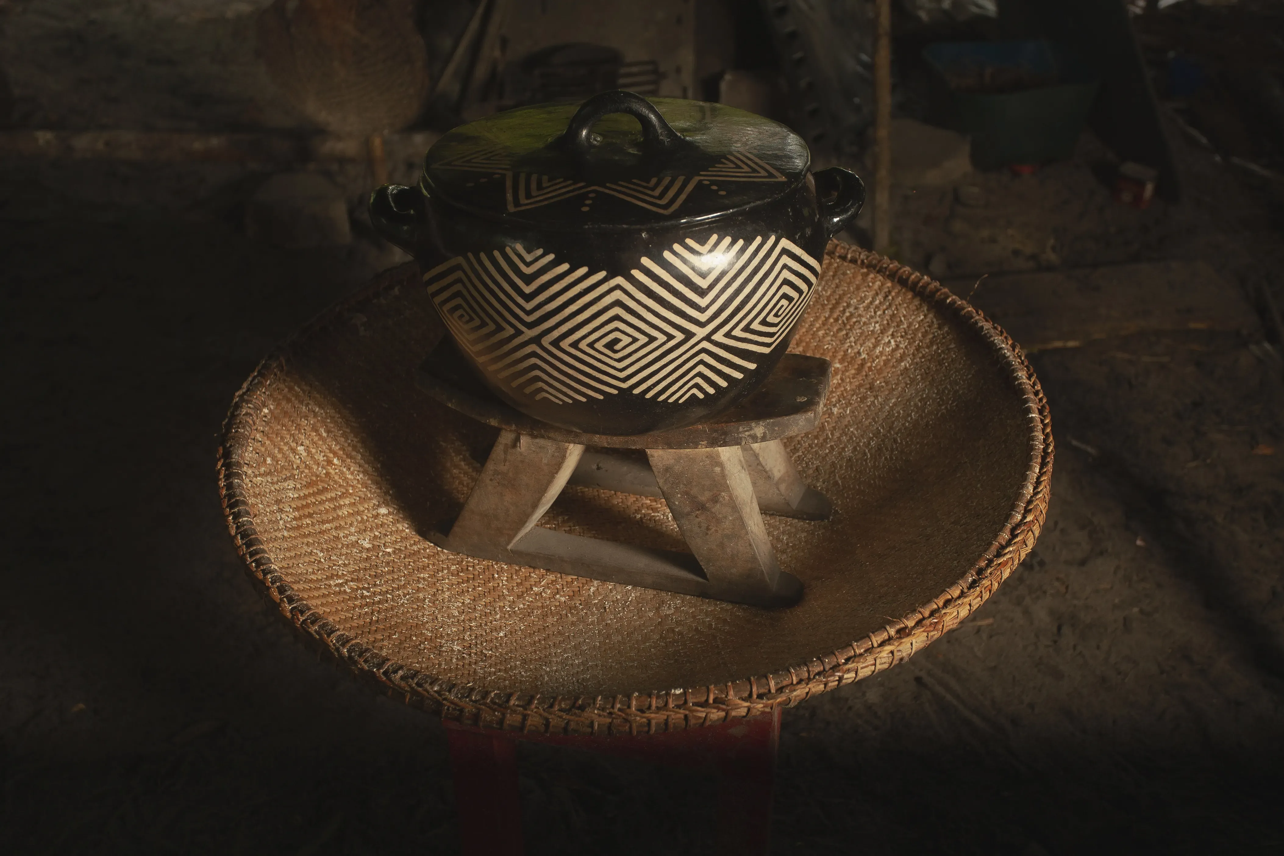 A piece of pottery sits on a small stool in a basket. The bowl is black with white lines going in parallel angles turning in on themselves, evoking a river current.