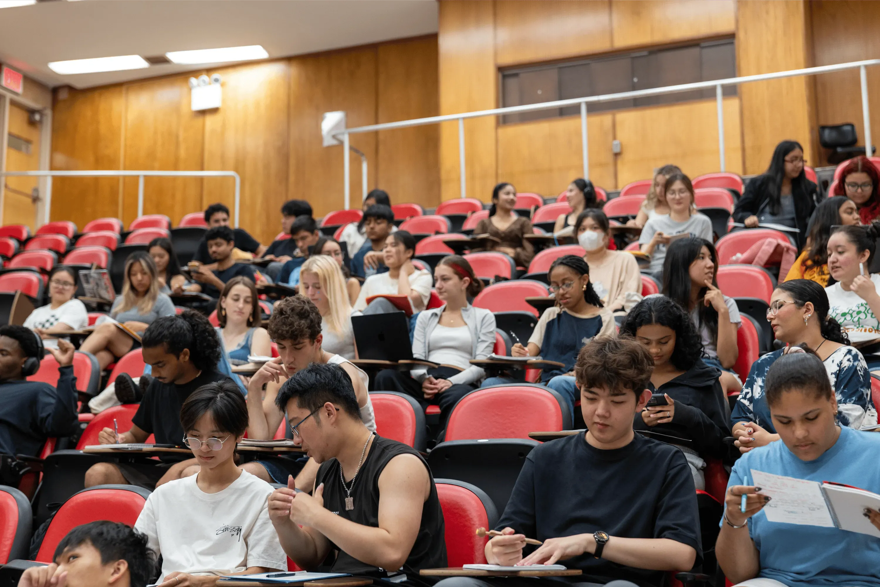 A photo of CUNY SEEK Program students in a lecture hall during class. A diverse group of students is spread across several rows of chairs. They have notetaking materials out and some appear to be in conversation with one another