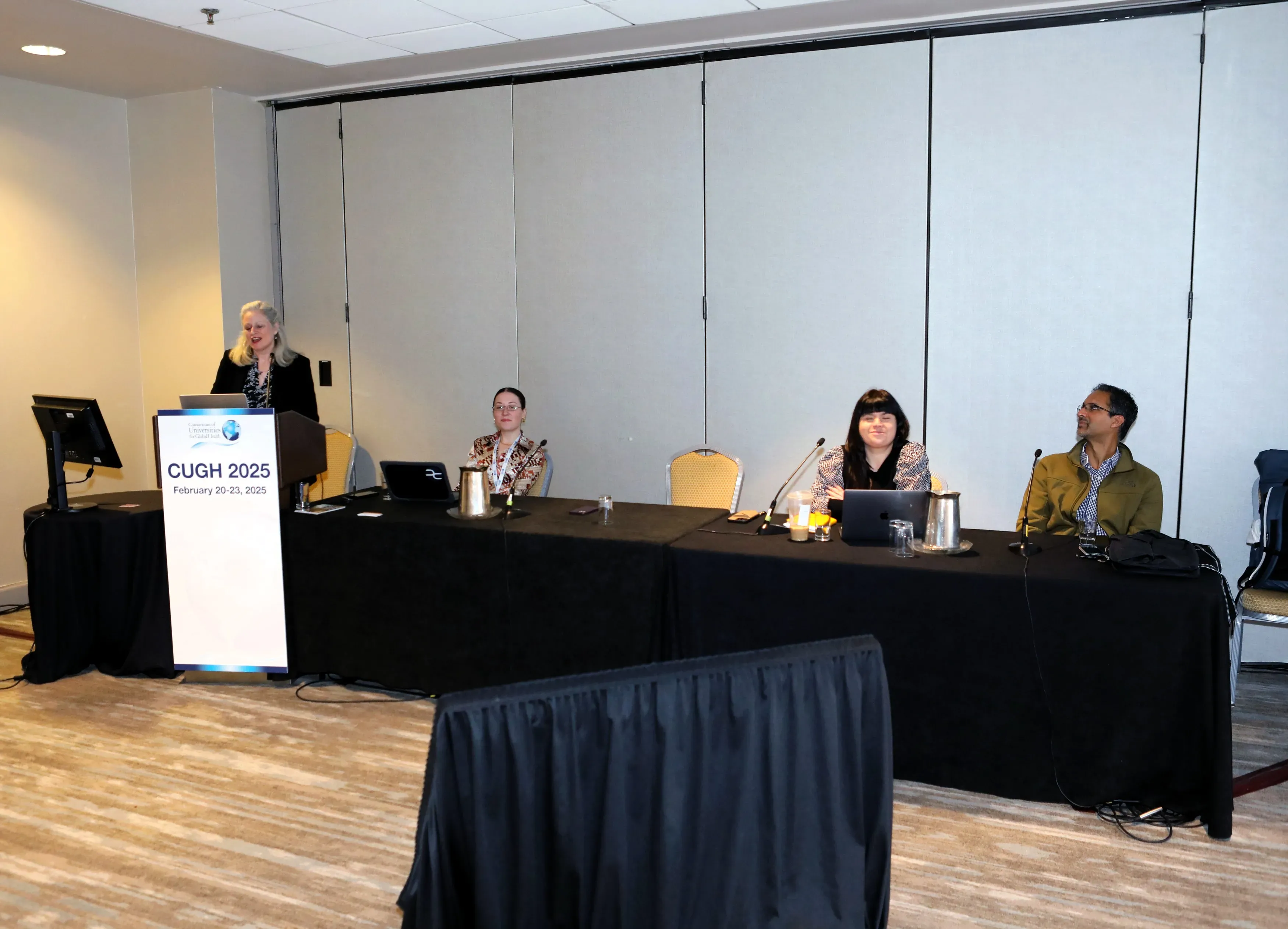 one person speaks at a podium and three people sit at a panel on global health and AI at CUGH Conference