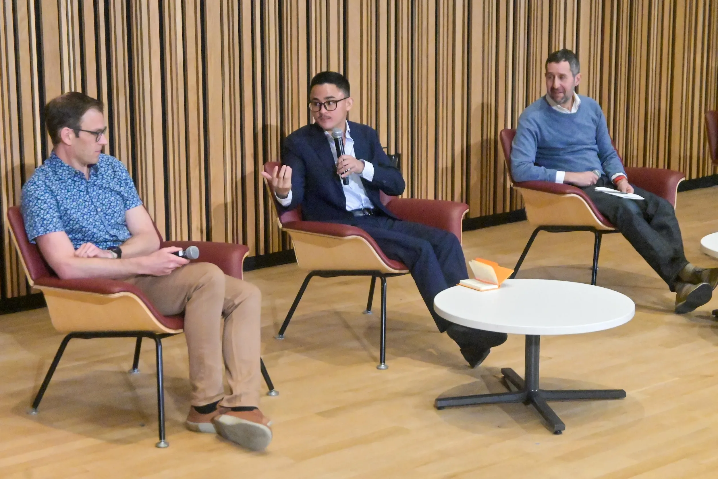Three men sit in chairs, one with microphone talks