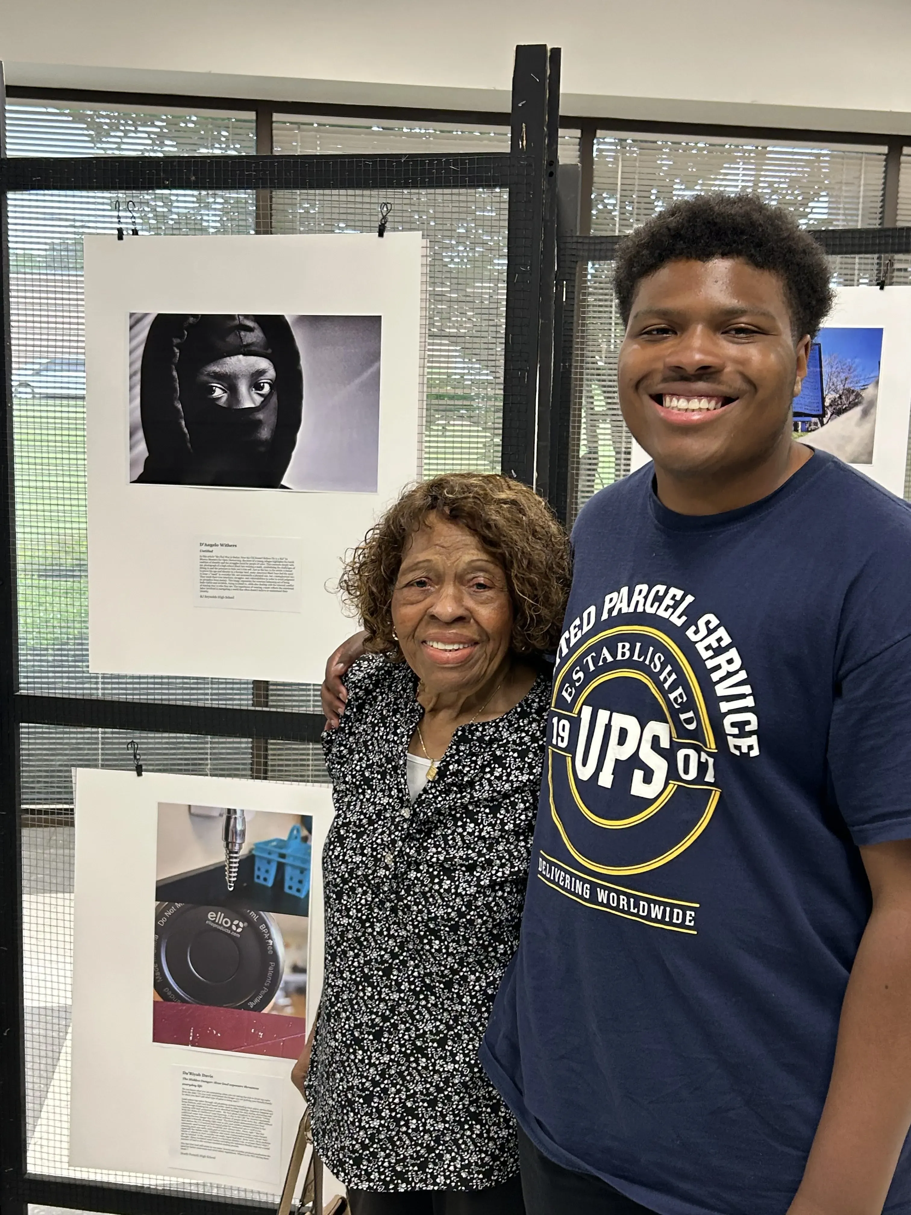 A student stands with an older woman next to the display of his photo from the "How I See It" photo contest. The photo is a black and white portrait of a young Black boy wearing a hoodie.