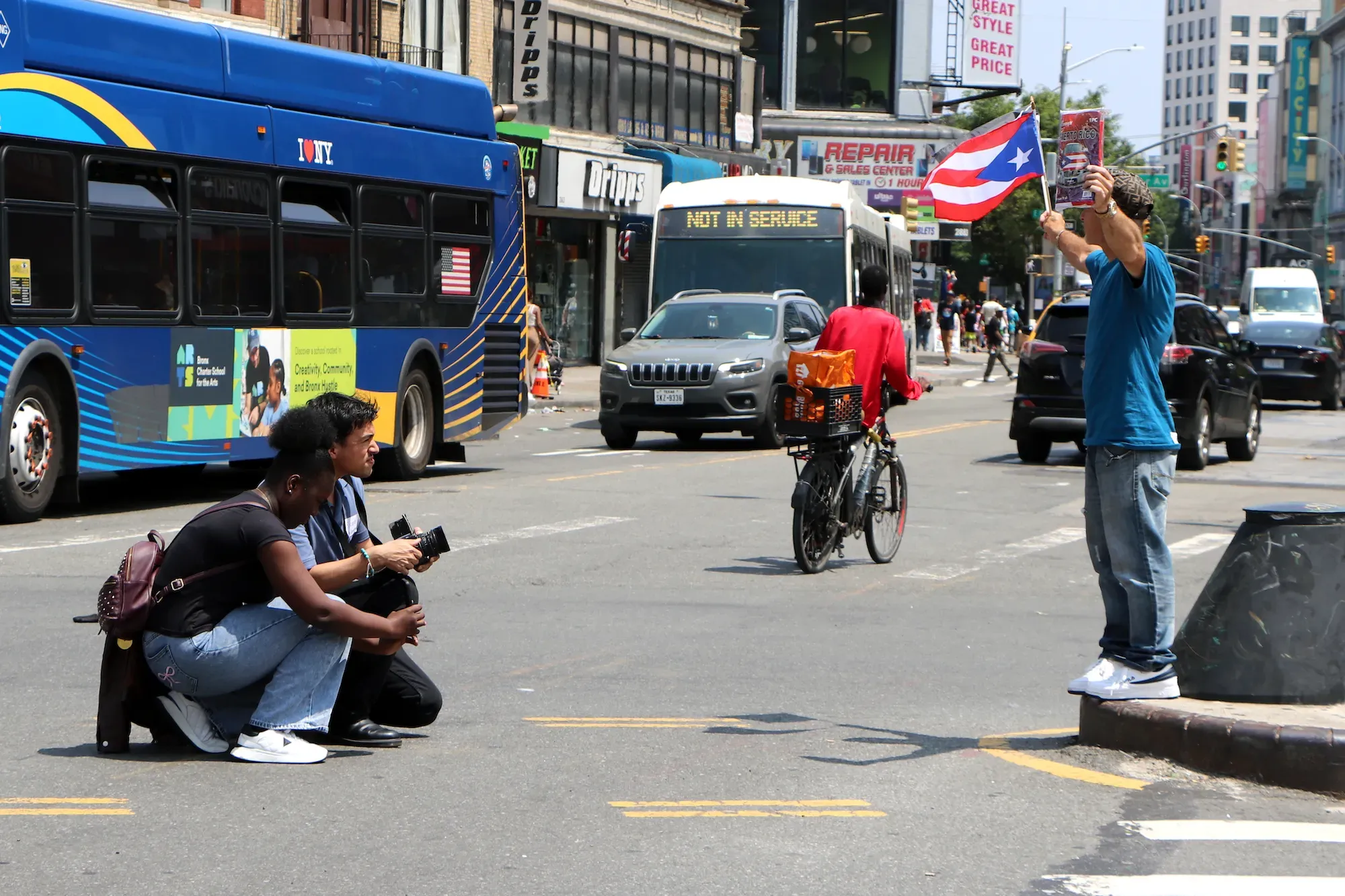 Two people kneel in the street and take a photo of a man holding flags