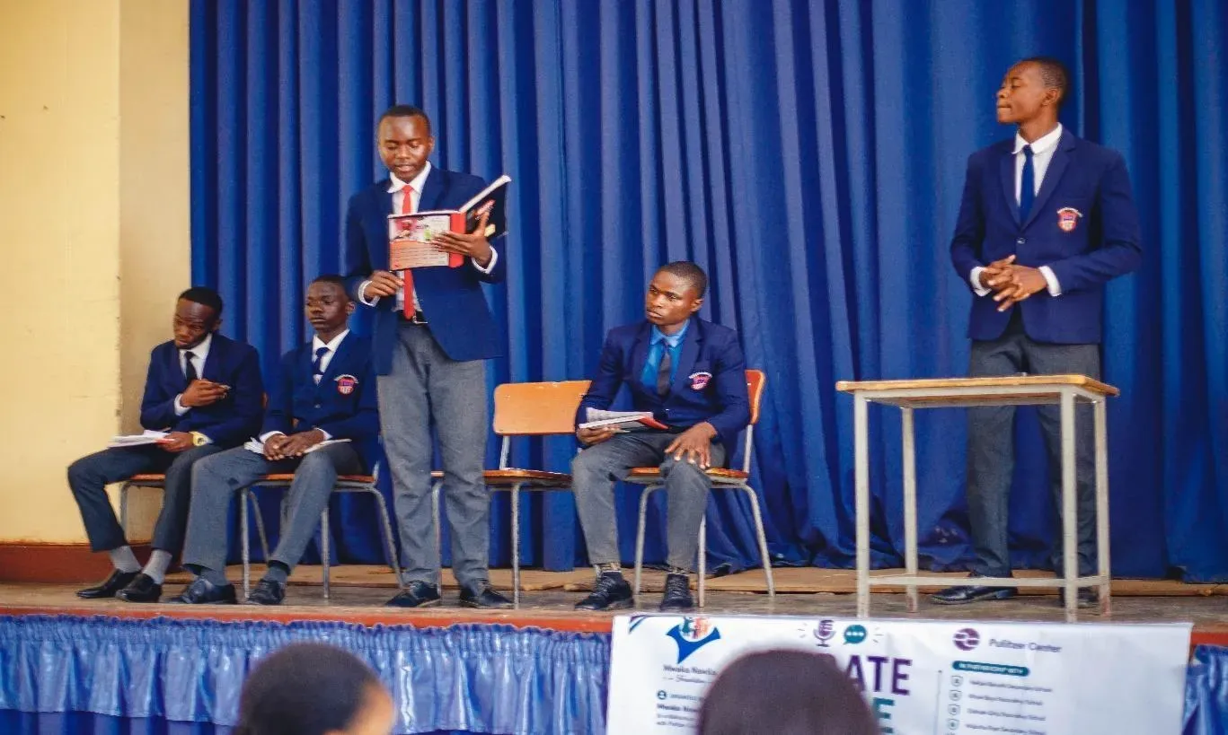 a student speaks on a stage. Another student stands listening, and three others sit behind them.