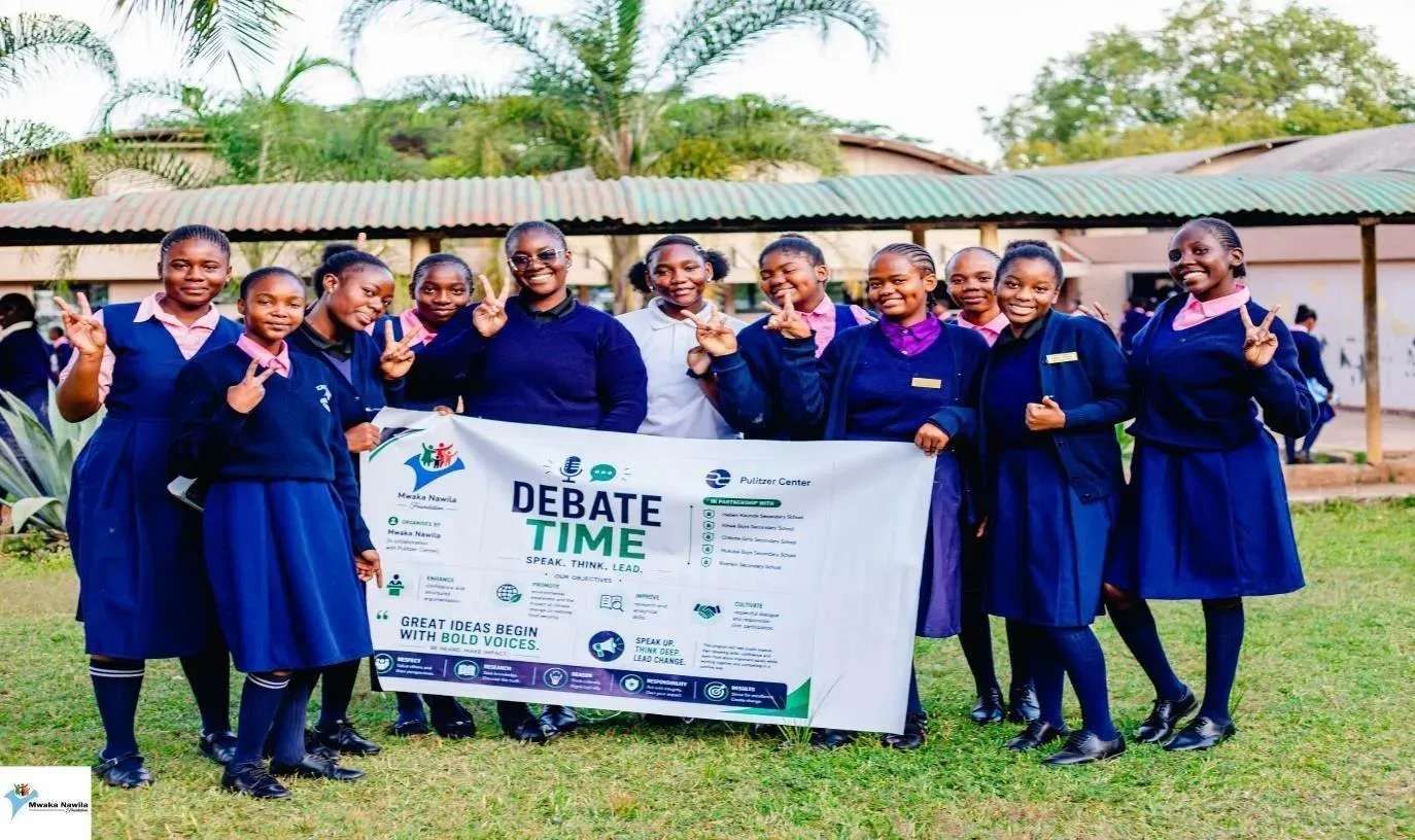 Schoolgirls pose for a group photo with a banner from the event titled 'debate time' with information about the sponsors.
