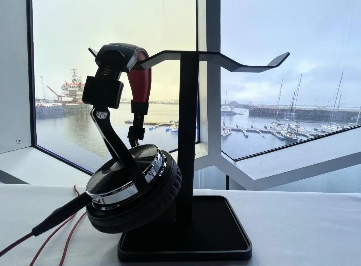 A pair of headphones hangs on a stand on a table, behind which is a large window showing a harbor