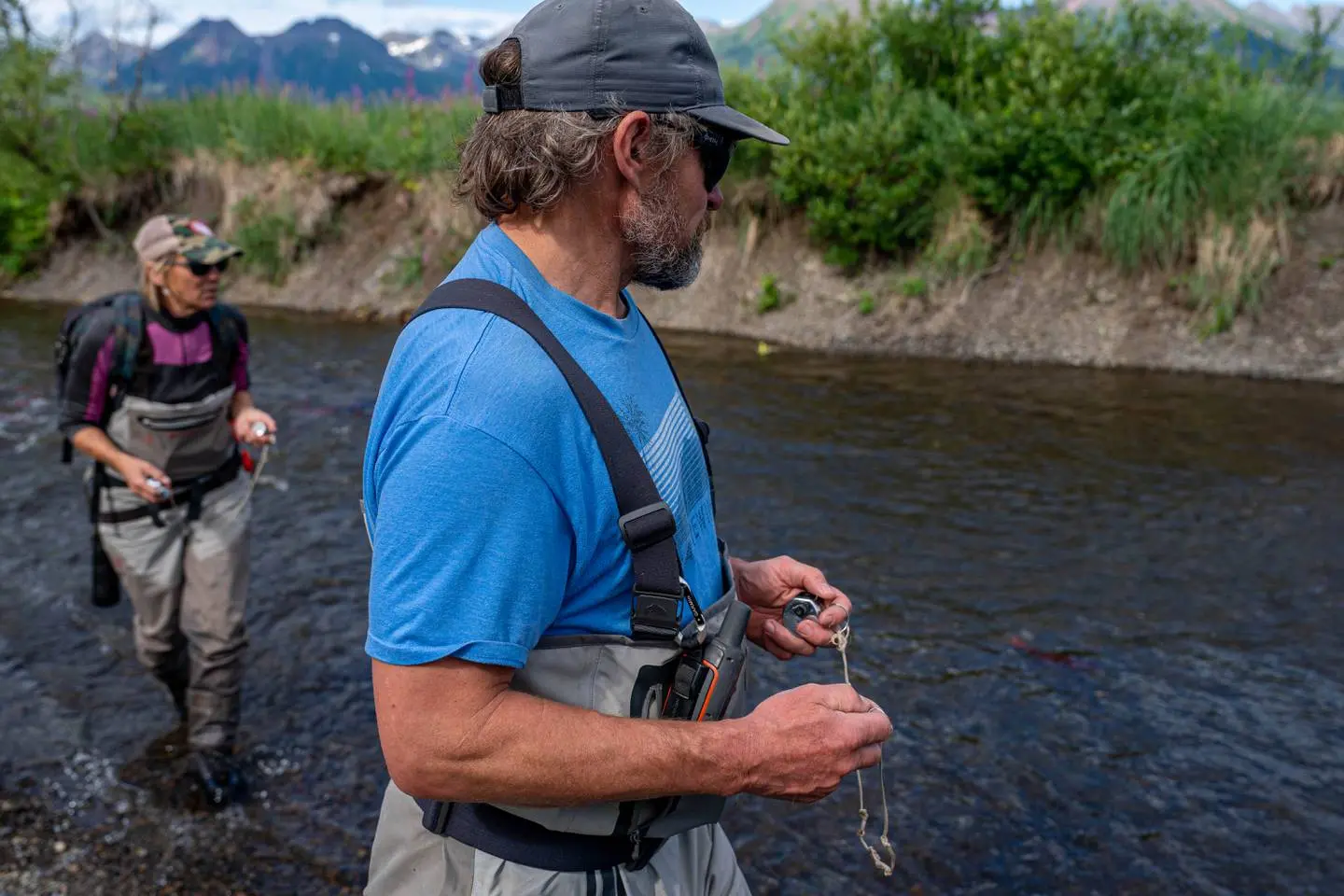 Two scientists wade in a creek. They both hold small metal counters in their hands as they scan the water. 